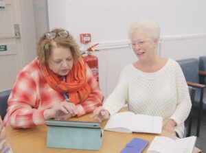 Two women are looking at a tablet, one is taking notes