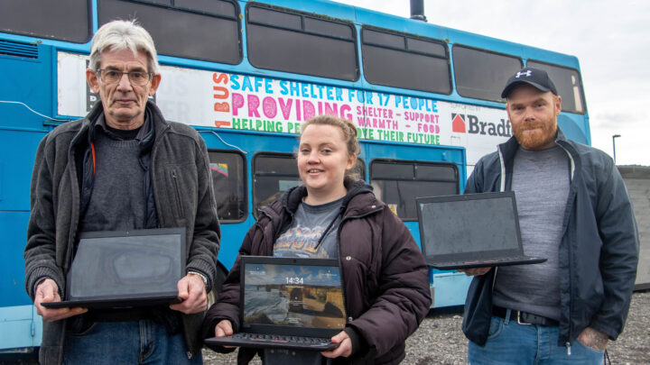Two men and a woman holding laptops they were gifted from Dorset Council's Digital Doorway scheme. They are in front of a double decker bus at the Bus Shelter site for homeless people in Weymouth.