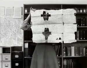 Black and white image of woman holding up an old document and peeking through a hole in it
