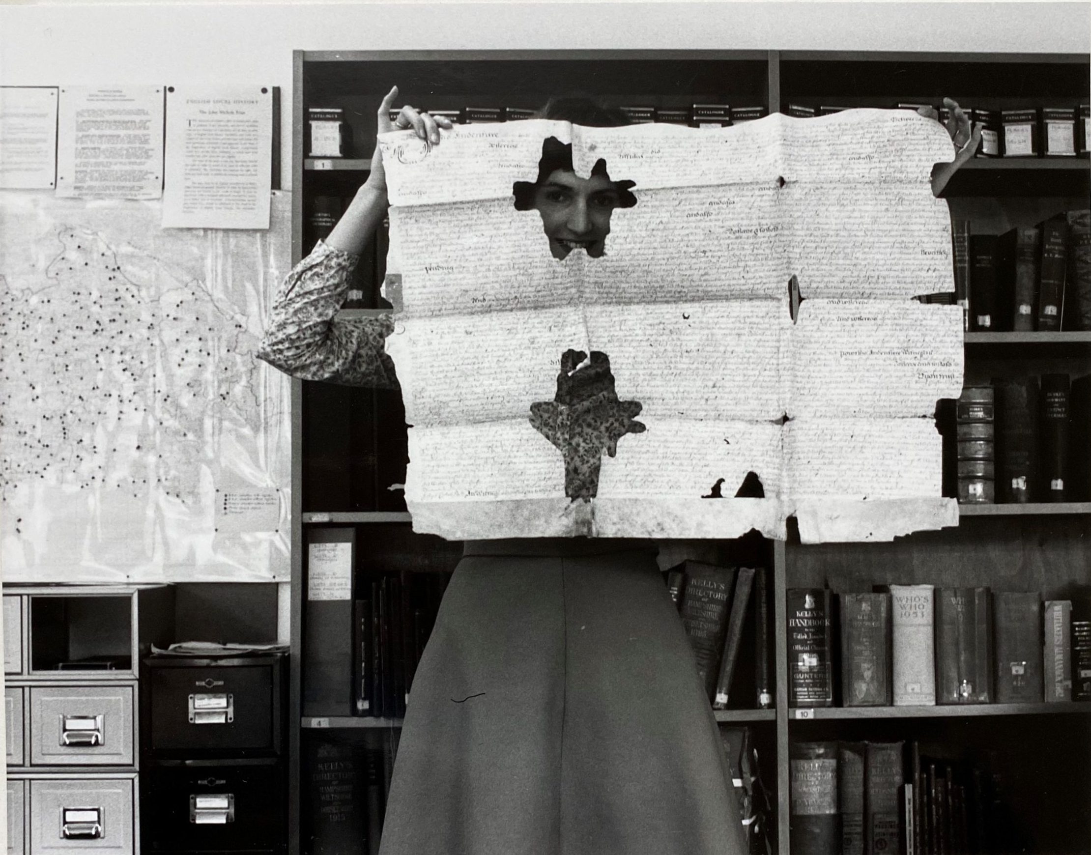 Black and white image of woman holding up an old document and peeking through a hole in it