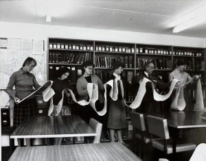 Black and white photograph of 6 people holding a very long archive document