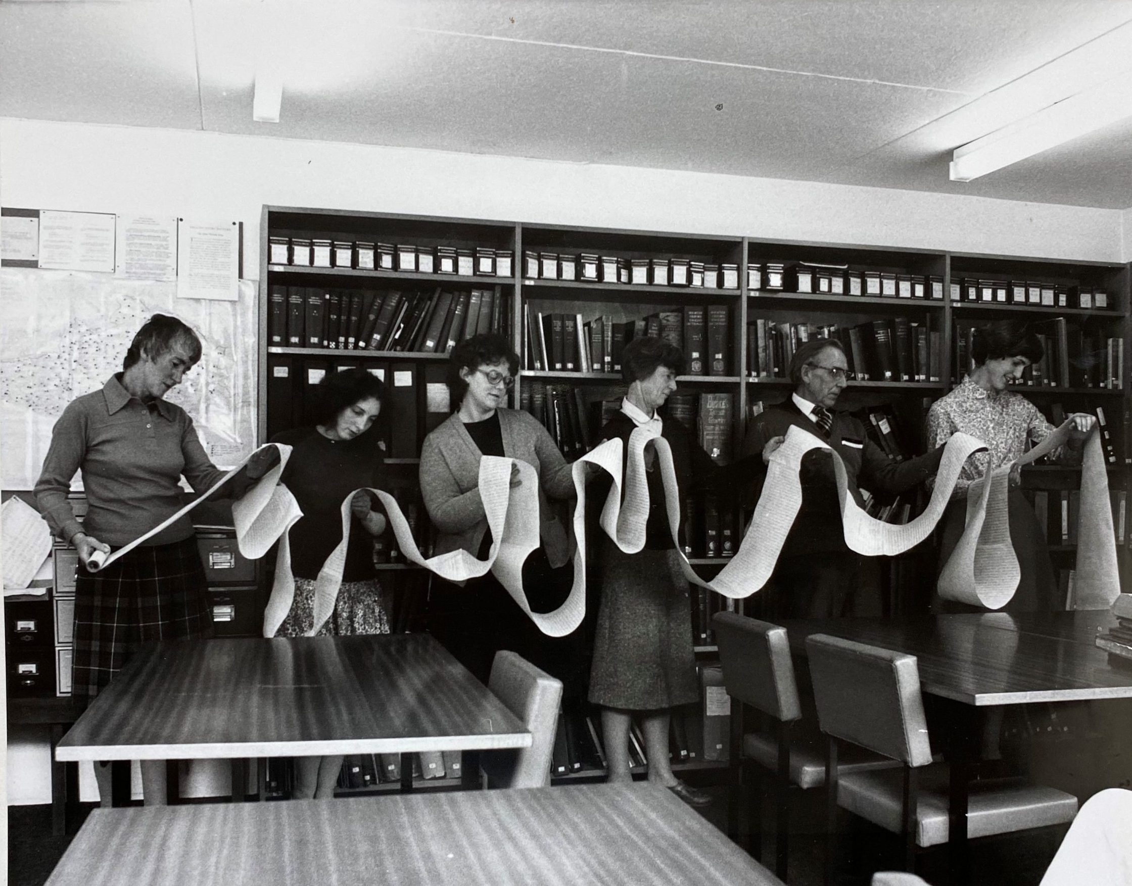 Black and white photograph of 6 people holding a very long archive document