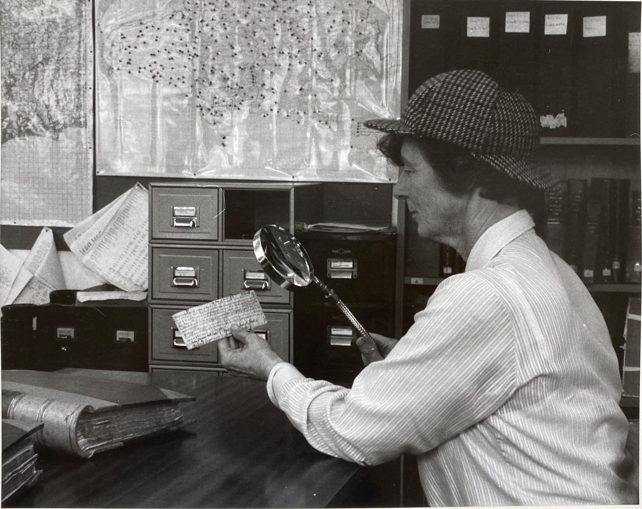 A black and white photograph of a woman wearing a deer stalker hat looking at a small document through a magnifying glass