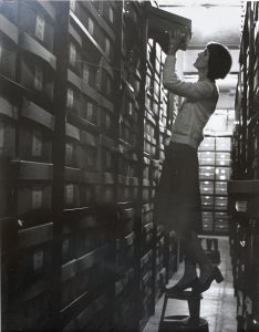Black and white photograph of a woman standing on a stool in the archive repository reaching for a box.