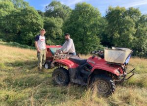 two individuals standing in field next to quad bike and farming machinery