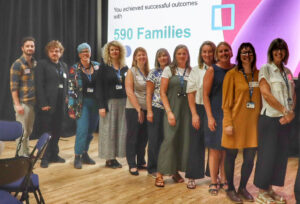 Group of adults stand in line and pose in front of screen which reads 'You achieved positive outcomes with 590 families'