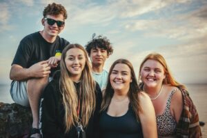five teens are smiling at the camera. The sea can be seen in the background