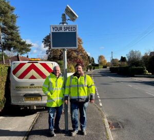 Two men wearing yellow jackets standing in front of a device that displays vehicle speeds